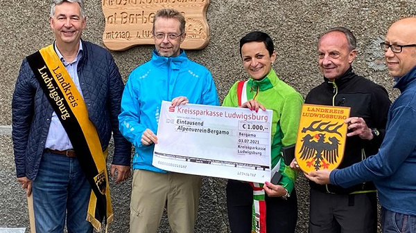 Cinq personnes se tiennent devant une plaque commémorative, tenant un chèque géant de 1000 euros. L’atmosphère est festive, avec des sourires et des vêtements variés. Elles représentent une donation au comité Alpeverein Bergmoos, symbolisant un soutien à une initiative locale.