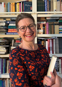 Eine lächelnde Frau mit Brille trägt ein blühendes, dunkles Kleid und hält ein Buch in der Hand. Im Hintergrund ist ein Bücherregal mit zahlreichen Büchern sichtbar.