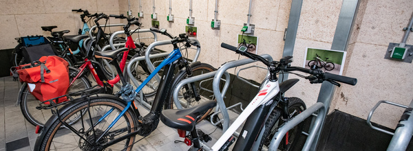Une salle de stationnement à vélos avec plusieurs vélos attachés à des supports. Certains vélos sont équipés de sacs. Des dispositifs de recharge sont visibles sur le mur. L'environnement est sobre et bien organisé, adapté aux cyclistes.