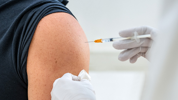 Un operatore sanitario somministra un vaccino a un paziente, visibile dal braccio scoperto. La persona indossa una maglietta scura e l'operatore tiene una siringa con una soluzione al suo fianco, pronto a effettuare l'iniezione.