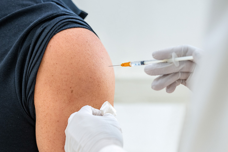 Eine Person erhält eine Impfung in den Oberarm. Eine Hand hält eine Spritze, während eine andere Hand ein Tuch bereitstellt. Der Arm ist gut sichtbar, und die Person trägt ein schwarzes T-Shirt.