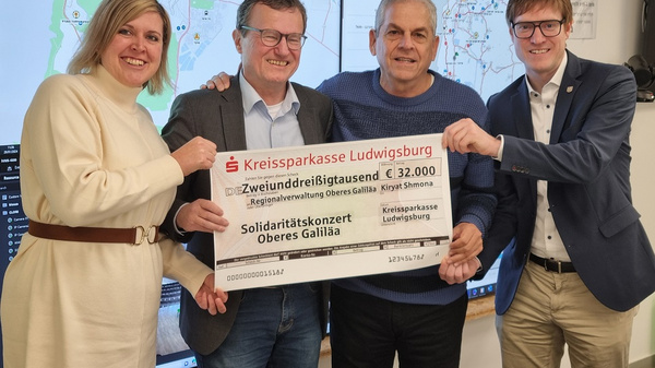 Vier Personen stehen zusammen und halten einen großen Scheck über 32.000 Euro in der Hand. Der Scheck ist an die "Regionalverwaltung Oberes Galiläa" adressiert und unterstützt ein Solidaritätskonzert. Im Hintergrund sind Bildschirme mit Karten und Informationen zu sehen.