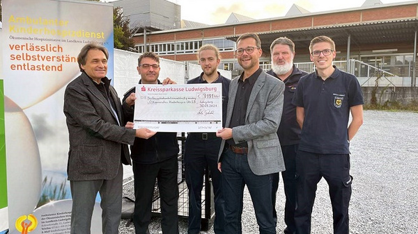 Un groupe de cinq personnes se tient devant un panneau d'affichage, tenant un grand chèque symbolique de la Kreissparkasse Ludwigsburg. Elles semblent célébrer une contribution financière, avec un fond d'établissement visible derrière elles.