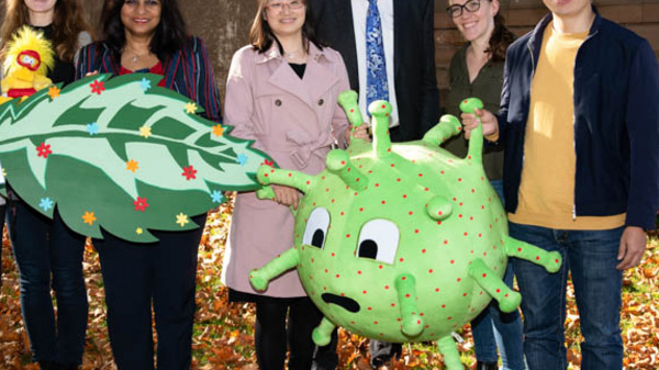 A group of eight people stands outdoors among fallen leaves. They are holding oversized cartoonish leaf and virus props, showcasing a fun and educational theme. The setting suggests a celebration or event related to science or ecology.