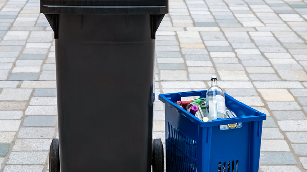 Ein schwarzer Müllcontainer mit blauer Klappe steht neben einem blauen Sammelkorb, der mit Flaschen und anderen recycelbaren Materialien gefüllt ist. Der Hintergrund besteht aus grauen Pflastersteinen.
