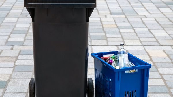 Ein schwarzer Müllcontainer mit blauer Klappe steht neben einem blauen Sammelkorb, der mit Flaschen und anderen recycelbaren Materialien gefüllt ist. Der Hintergrund besteht aus grauen Pflastersteinen.