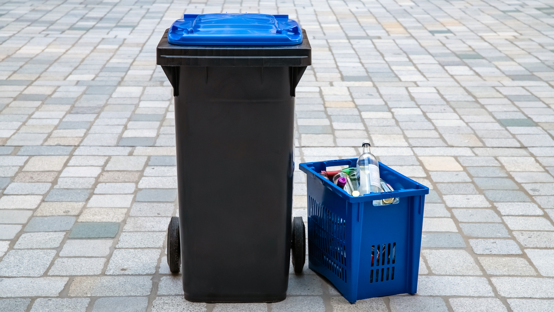 A black trash bin with a blue lid stands next to a blue recycling bin filled with various recyclable items, including a plastic bottle. They are placed on a patterned stone pavement.