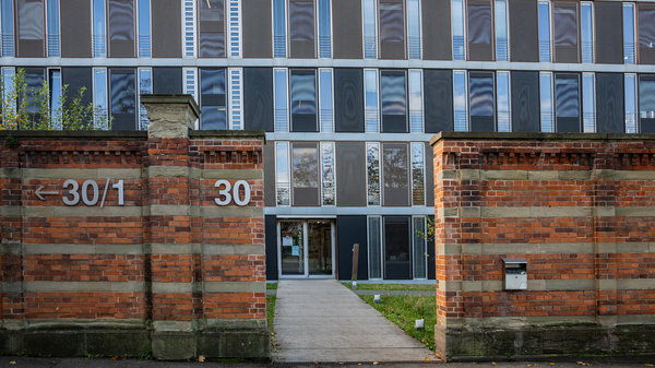 A modern building with large glass windows is set behind a brick wall. The entrance, flanked by red bricks, features door numbers "30" and "30/1". A pathway leads to the door, with green grass visible on one side.