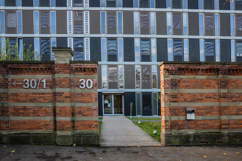 Une entrée principale d'un bâtiment moderne avec une façade vitrée, entourée de murs en briques anciennes. Le numéro 30 est visible, ainsi qu'une indication vers le côté gauche. L'espace est bien dégagé avec une petite pelouse à l'avant.