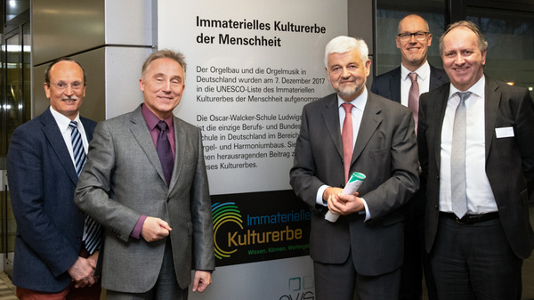 Five men standing together in front of a large sign that reads "Immaterielle Kulturerbe der Menschheit." The men are dressed in formal attire, smiling, and appear to be engaging in a discussion or celebration regarding cultural heritage recognition.
