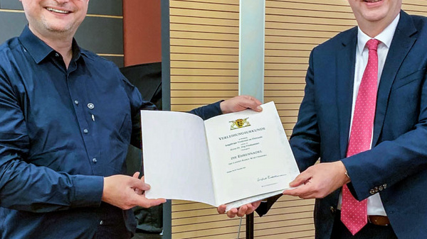 Zwei Männer stehen auf einer Bühne. Der linke Mann trägt ein schwarzes Hemd und lächelt, während er eine Urkunde in der Hand hält. Der rechte Mann trägt einen Anzug und präsentiert ebenfalls die Urkunde. Beide schauen lächelnd in Richtung Kamera.