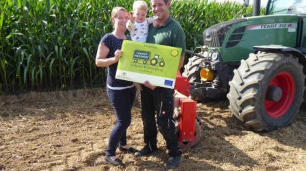 A smiling family stands in a field, with a corn crop in the background. They hold a large sign promoting agricultural equipment, while a tractor is parked nearby. The family includes a woman, a man, and a small child.