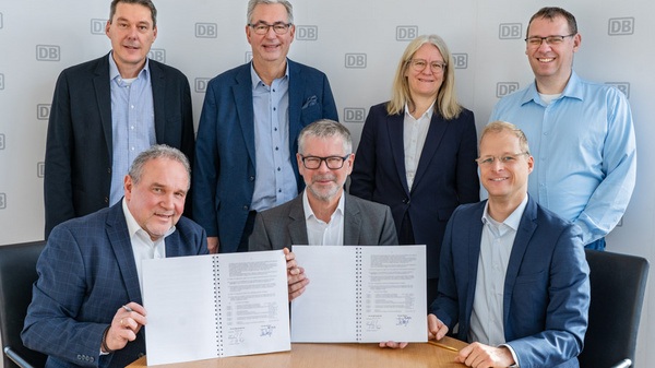 Eine Gruppe von sieben Personen steht und sitzt um einen Tisch. Zwei Männer in der Vordergrund halten ein Dokument mit Unterschriften. Im Hintergrund sind weitere Personen, die in gleicherweise für die Aufnahme posieren. Das Umfeld scheint eine Büroumgebung zu sein.