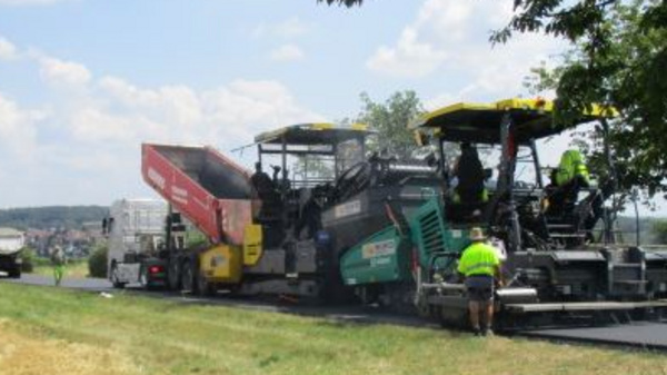A road construction scene shows workers operating heavy machinery, including a paver and a truck, alongside an open field. The sky is clear with a few clouds, and trees are in the background. The focus is on the construction activity and the environment.