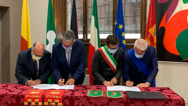 Four individuals are gathered around a table, signing documents. They are wearing masks, and two of them are in formal attire, while the third is wearing a mayor's sash. The setting includes flags and a colorful abstract painting in the background. The documents and table have decorative elements.
