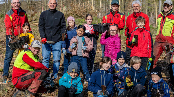 Eine Gruppe von Kindern und Erwachsenen steht im Wald und hält Setzlinge in der Hand. Sie lächeln und posieren gemeinsam für ein Foto, während einige in Arbeitshosen und -jacken gekleidet sind. Die Szene vermittelt ein Gefühl von Gemeinschaft und Engagement für das Pflanzen von Bäumen.