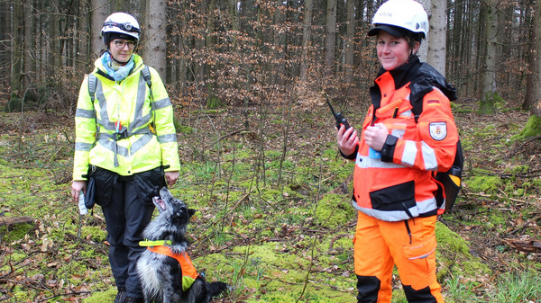 Zwei Rettungskräfte stehen im Wald. Eine Person trägt eine gelbe Warnjacke und hält ein Gerät in der Hand. Neben ihr sitzt ein Hund. Die andere Person ist in orangefarbener Kleidung gekleidet und hält ebenfalls ein Gerät. Beide blicken in die Kamera.