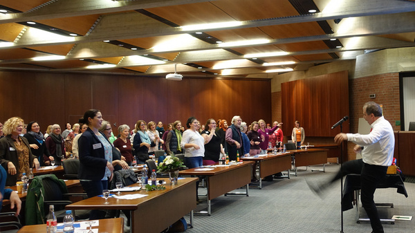 A crowd of people stands attentively in a conference room, facing a speaker who is performing animatedly. Tables are set with papers and bottles. The atmosphere appears lively and engaged, with the audience actively participating in the presentation.
