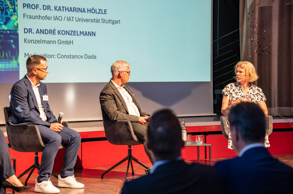 Drei Personen sitzen auf Stühlen auf einer Bühne während einer Diskussionsrunde. Eine Frau moderiert das Gespräch, während zwei Männer neben ihr sitzen. Im Hintergrund sind Präsentationsfolien sichtbar. Die Atmosphäre ist professionell und auf Interaktion ausgelegt.