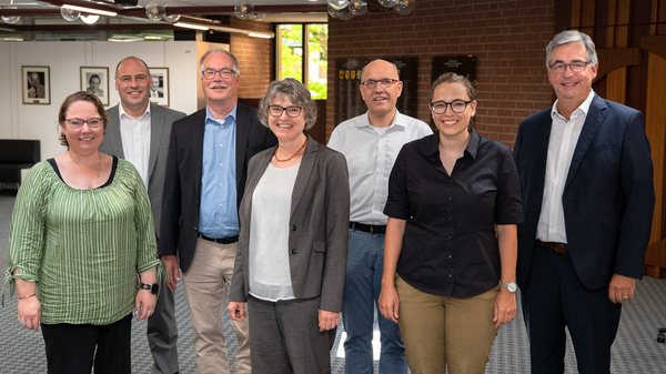 Eine Gruppe von sieben Personen steht in einem Raum. Sie sind in formeller Kleidung und lächeln. Im Hintergrund sieht man eine moderne Raumgestaltung mit Fenstern und beleuchteten Elementen. Die Personen scheinen eine positive, kollegiale Stimmung auszustrahlen.