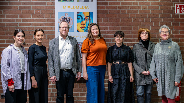 Eine Gruppe von sieben Personen steht vor einer Wand mit einem Plakat, das "MIXEDMEDIA" zeigt. Die Frauen und Männer sind unterschiedlich gekleidet und lächeln in die Kamera. Die Umgebung wirkt freundlich und einladend.
