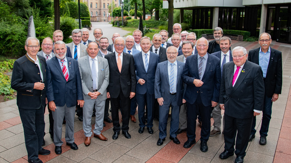 A group of around 30 men stands together outdoors in a courtyard, smiling and dressed in formal attire. The background features greenery and buildings, indicating a professional gathering or event. The atmosphere appears friendly and welcoming.