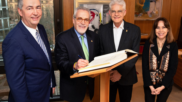Four individuals stand together in a well-lit room. One person is signing a document on a podium, while the others watch with smiles. The setting features wooden walls and abstract artwork, suggesting a formal or celebratory occasion.