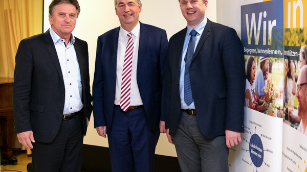 Three men stand together in front of a presentation backdrop. They are smiling and dressed in professional attire. The backdrop features the text "Wir in City" and images promoting community engagement and quality of life. The setting appears to be a formal event or meeting.