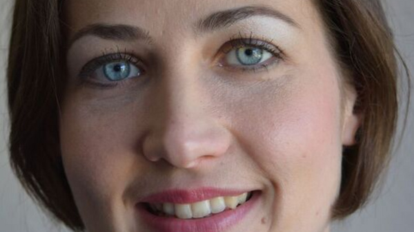 Ein freundliches Porträt einer Frau mit kurzen, braunen Haaren und blauen Augen. Sie trägt ein schwarzes Oberteil und lächelt. Der Hintergrund ist neutral und hell, wodurch der Fokus auf ihrem Gesicht liegt.