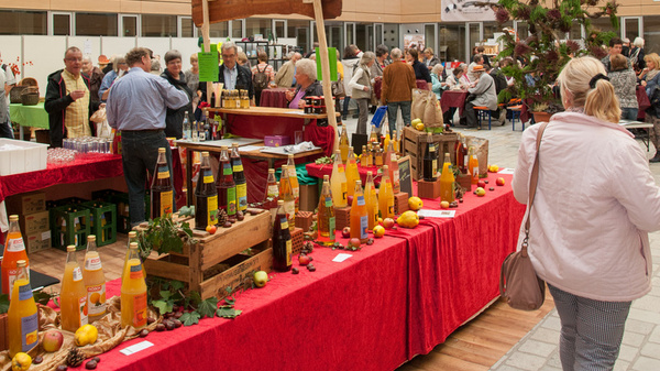 Un espace intérieur animé avec des tables recouvertes de nappes rouges présentant diverses boissons. Des personnes discutent et se déplacent dans l'aire, entourées de grandes fenêtres laissant entrer la lumière. L'atmosphère est conviviale, indiquant un événement festif.