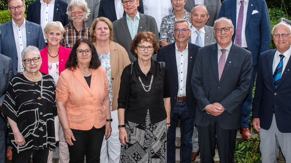 Eine Gruppe von 20 Personen steht im Freien in einer Gartenumgebung. Die Menschen sind in formeller Kleidung gekleidet, und einige lächeln in Richtung der Kamera. Im Hintergrund sind Büsche und ein Gebäude zu sehen.