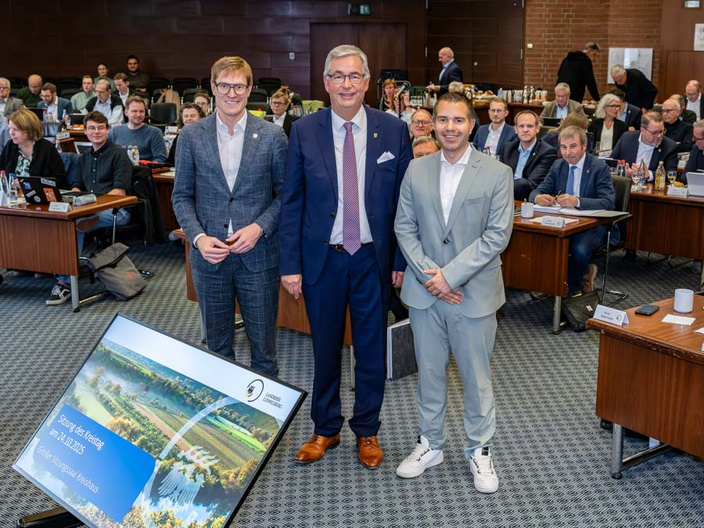 Drei Männer stehen in einem Konferenzraum. Sie lächeln in die Kamera, während viele Menschen im Hintergrund sitzen und an Tischen arbeiten. Ein Bildschirm im Vordergrund zeigt eine Landschaft mit dem Titel "Wasser der Zukunft".