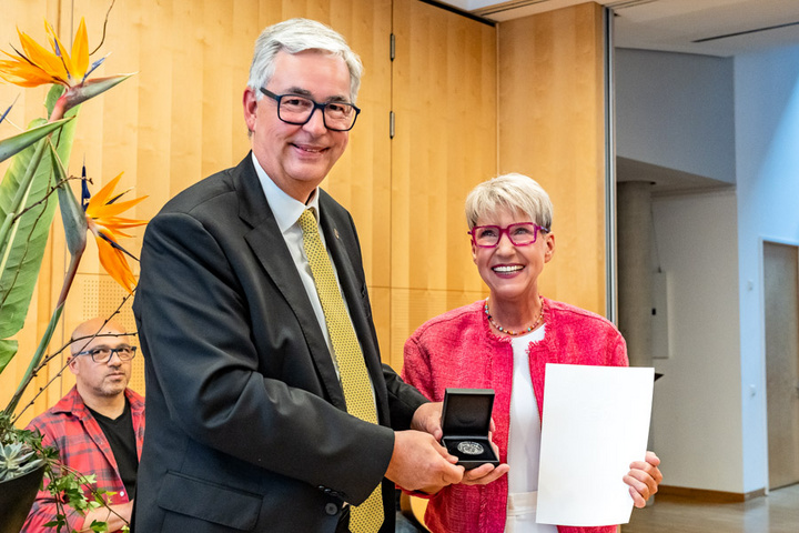Eine Person im Anzug überreicht einer Frau mit Brille eine Auszeichnung in einer kleinen Schachtel, während sie ein Zertifikat in der anderen Hand hält. Im Hintergrund sind unscharfe Silhouetten von weiteren Personen und eine Pflanze zu sehen.