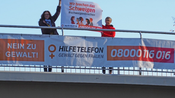 Deux personnes se tiennent sur un pont, tenant une grande affiche où l'on peut lire "Nous brisons le silence". En dessous, une bannière indique "Non à la violence !" et un numéro d'aide pour les femmes victimes de violence.