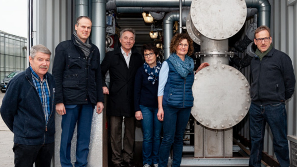 Un gruppo di sei persone si trova davanti a un contenitore industriale. All'interno si possono vedere tubi e un grande serbatoio cilindrico. I membri del gruppo indossano abbigliamento casual e si posizionano in varie pose, sorridendo verso la fotocamera.