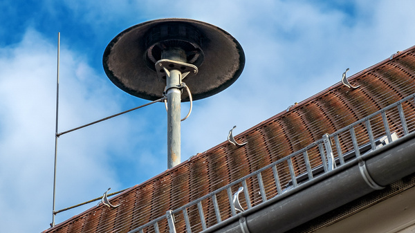 Ein Lautsprecher auf dem Dach eines Gebäudes, vor einem blauen Himmel mit wenigen Wolken. Der Lautsprecher hat eine runde Form und ist an einem speziellen Mast montiert.