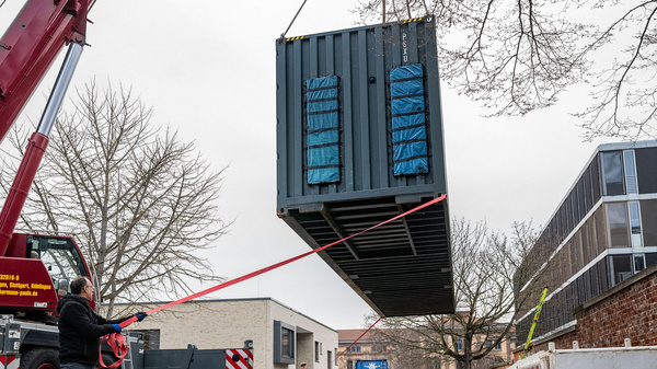 Ein Kran hebt einen Baucontainer an, während ein Arbeiter ihn mit einem Seil führt. Im Hintergrund sind Bäume und Gebäude sichtbar. Die Szene zeigt eine Baustelle, an der gerade Arbeiten durchgeführt werden.