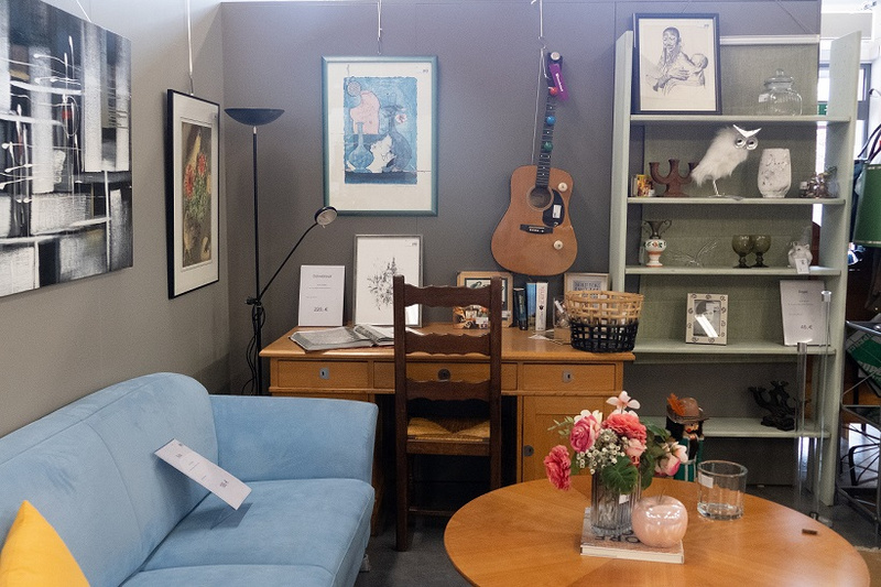 Une pièce de mobilier présentée avec un canapé bleu, une table ronde avec des fleurs, une étagère contenant divers objets, et un bureau en bois. Des œuvres d'art sont accrochées aux murs, et une guitare est suspendue sur le côté.