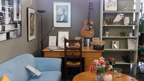 Ein gemütliches Zimmer mit einem blauen Sofa, einem Holztisch und einem Stuhl. Auf dem Tisch steht eine Vase mit rosa Blumen. An der Wand hängen Bilder, und ein Regal mit Büchern und Deko ist sichtbar. Ein Akustikgitarre hängt an der Wand.