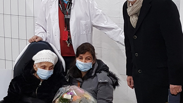 A medical professional in a white coat stands behind two women seated in a medical setting. One woman, wearing a hospital beanie, holds a bouquet of flowers, while the other woman, in a gray jacket, smiles. They all wear face masks. A sign on the wall indicates "8."