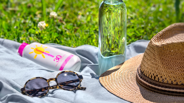 Ein Handtuch liegt auf grünem Gras, darauf befinden sich eine Flasche Wasser, eine Sonnencreme mit gelbem Etikett und eine Sonnenbrille. Neben diesen Gegenständen liegt ein Strohhut. Die Szene vermittelt ein entspanntes Sommergefühl.