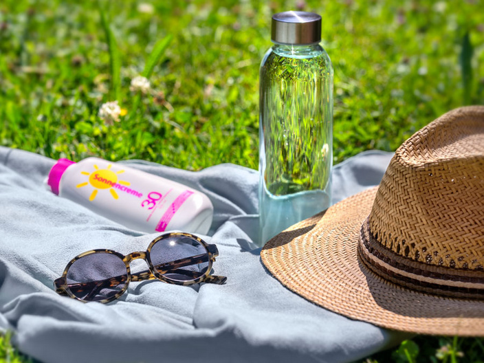 Ein Handtuch liegt auf grünem Gras, darauf befinden sich eine Flasche Wasser, eine Sonnencreme mit gelbem Etikett und eine Sonnenbrille. Neben diesen Gegenständen liegt ein Strohhut. Die Szene vermittelt ein entspanntes Sommergefühl.