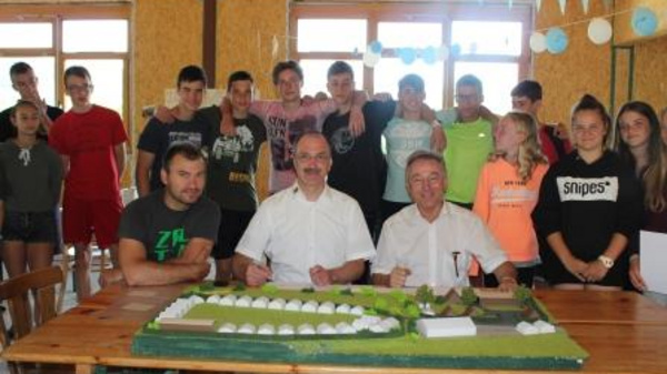 A group of students and two adults are gathered around a table showcasing a detailed model of a landscape. The adults are seated at the front, actively engaged with the model, while the students stand behind them, smiling and posing for the photo in a room decorated with simple party decor.