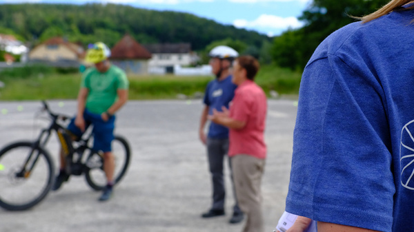 A group of people is gathered outdoors for an e-bike safety training session. One person in the foreground, wearing a blue shirt with a bicycle graphic, looks on while another person speaks. Several participants are on bicycles, and the setting is in a green, open area with trees in the background.