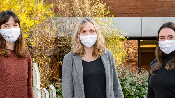 Three women stand outdoors, wearing white face masks. They are positioned in front of a brick building, amidst greenery and autumn foliage. The women are dressed in casual clothing, with one in a brown sweater, another in a gray cardigan, and the third in a black top.