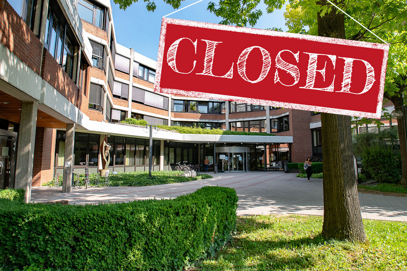 Das Bild zeigt einen Eingangsbereich eines Gebäudes mit einem deutlich sichtbaren Schild, auf dem "CLOSED" in großen roten Buchstaben steht. Die Umgebung ist hell und freundlich, mit Bäumen und gepflegten Grünflächen, aber das Gebäude ist geschlossen. (Text KI generiert)