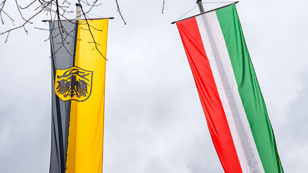 Zwei Fahnen wehen im Wind. Links die deutsche Flagge in Schwarz-Gelb mit dem Bundesadler, rechts die italienische Flagge in Grün, Weiß und Rot. Der Himmel ist bewölkt.