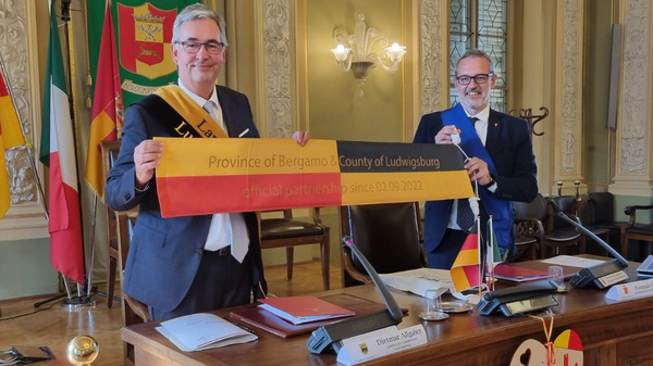 Two officials stand behind a wooden table in a formal room, holding a banner that reads "Province of Bergamo - County of Luxembourg" while presenting a partnership event. Flags of various nations are displayed in the background. The atmosphere is celebratory and diplomatic.