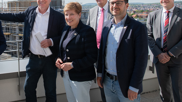 Eine Gruppe von sechs Personen steht auf einem Dach mit einer städtischen Landschaft im Hintergrund. Sie lächeln und posieren für das Foto. Der Himmel ist klar und sonnig, und im Hintergrund sind Bauarbeiten sichtbar. Es handelt sich um ein offizielles Treffen.