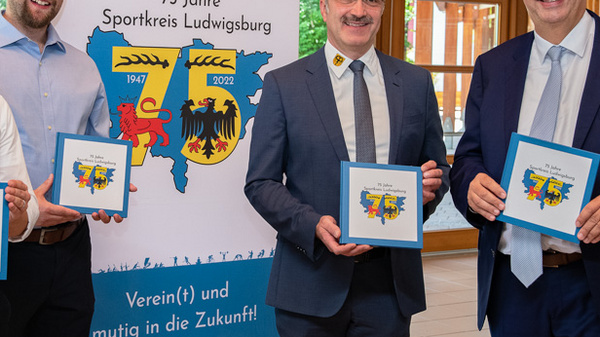 Vier Männer stehen nebeneinander und halten jeweils ein Buch in der Hand. Im Hintergrund ist ein Banner mit dem Text „75 Jahre Sportkreis Ludwigsburg“ und einem farbenfrohen Wappen zu sehen. Die Männer tragen formelle Kleidung und lächeln.